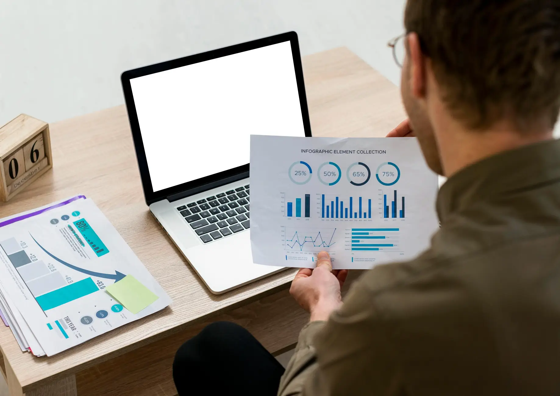 A man examining a report with graphs and data, sitting at a desk beside a laptop and a calendar displaying