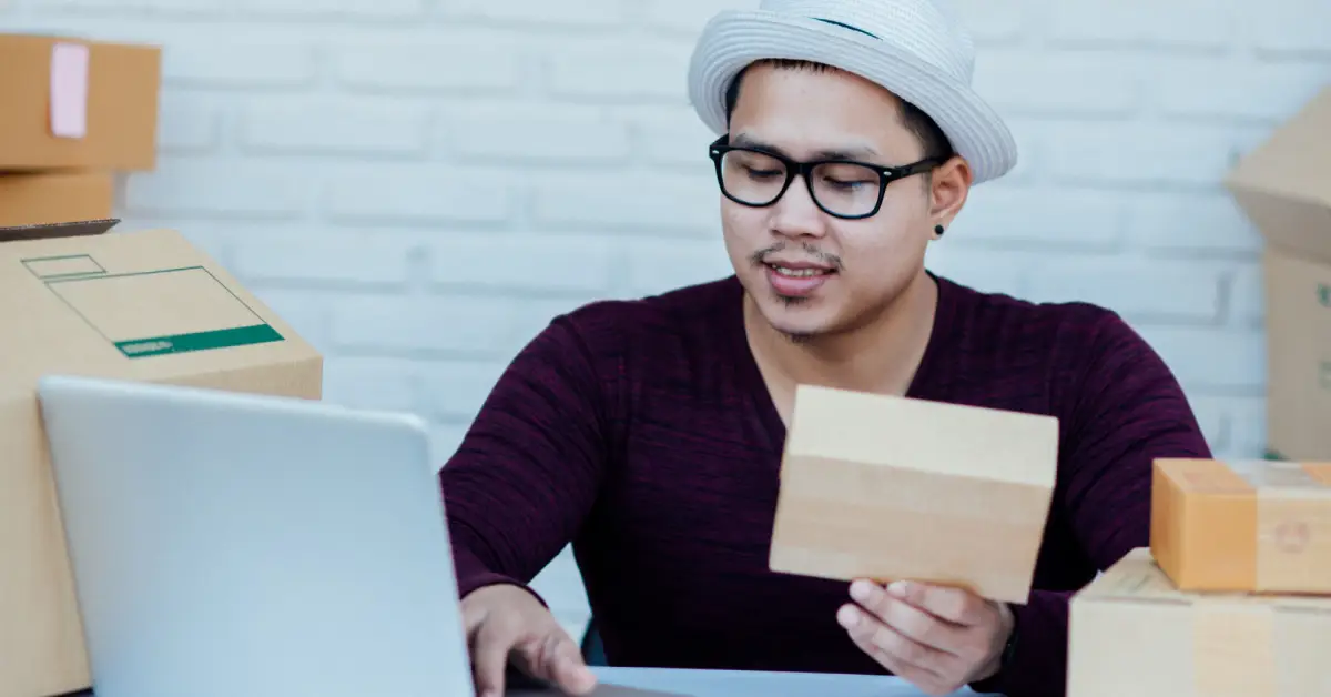 A person sits at a desk with a laptop, holding a small package, surrounded by various delivery boxes against a white brick wall A person sits at a desk with a laptop, holding a small package, surrounded by various delivery boxes against a white brick wall